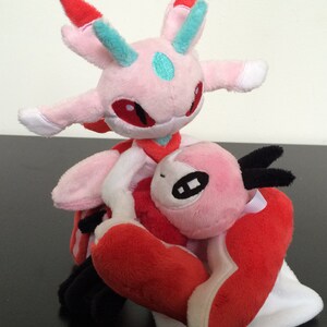 May include: Pink and red plush toy with a white belly and a black horn. The toy is sitting on top of another pink and red plush toy with a white belly and a blue horn.