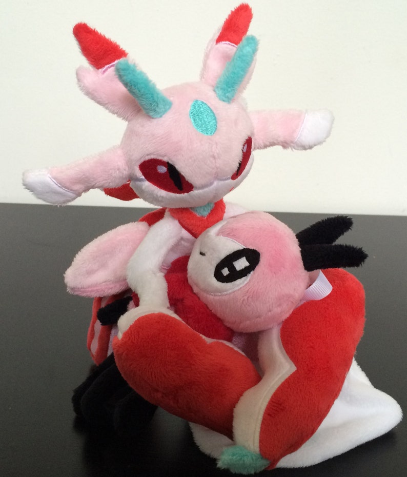 May include: Pink and red plush toy with a white belly and a black horn. The toy is sitting on top of another pink and red plush toy with a white belly and a blue horn.