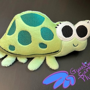 May include: A plush turtle toy in shades of green and yellow. The turtle has large, expressive eyes with black pupils and white outlines. The shell features darker green spots. The toy has a smiling face and soft, plush limbs. The text "Guardian Earth Plush" is visible.