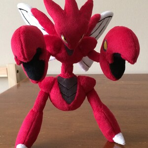 K&ouml;nnte beinhalten: Ein rotes und schwarzes Pl&uuml;schtier eines Pokemon-Charakters mit gro&szlig;en Krallen und Fl&uuml;geln. Das Spielzeug steht auf einer Holzoberfl&auml;che.