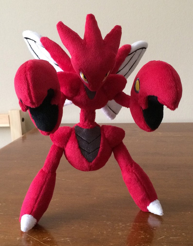 K&ouml;nnte beinhalten: Ein rotes und schwarzes Pl&uuml;schtier eines Pokemon-Charakters mit gro&szlig;en Krallen und Fl&uuml;geln. Das Spielzeug steht auf einer Holzoberfl&auml;che.