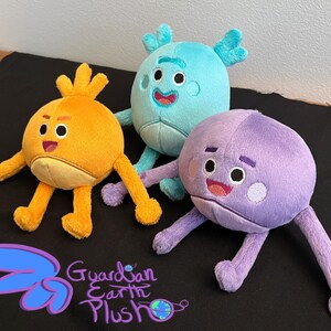 Puede incluir: Tres juguetes de peluche, uno naranja, uno azul y uno morado, con caras amigables y extremidades largas. Los juguetes est&aacute;n sentados sobre una superficie negra. El juguete naranja tiene un peinado naranja puntiagudo. El juguete azul tiene cuernos. El juguete morado tiene una amplia sonrisa. Los juguetes miden aproximadamente 25 cm de alto.