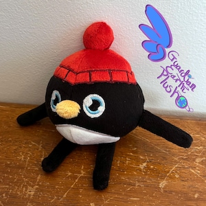 Puede incluir: Un juguete de peluche de ping&uuml;ino blanco y negro que lleva un gorro de punto rojo con un pomp&oacute;n blanco. El ping&uuml;ino tiene ojos bordados y un pico amarillo.