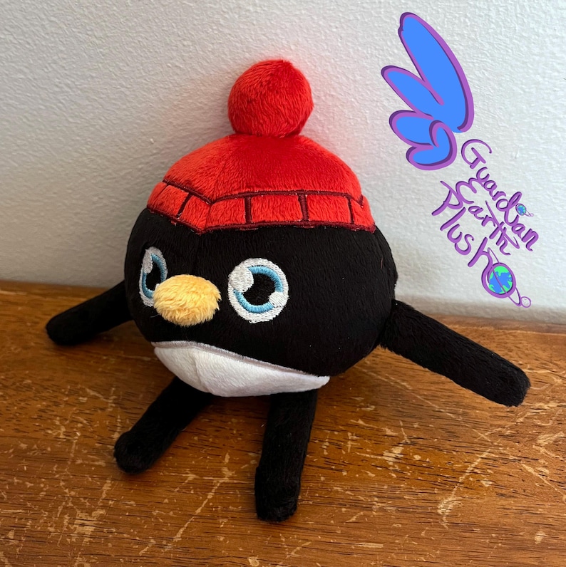 Puede incluir: Un juguete de peluche de ping&uuml;ino blanco y negro que lleva un gorro de punto rojo con un pomp&oacute;n blanco. El ping&uuml;ino tiene ojos bordados y un pico amarillo.