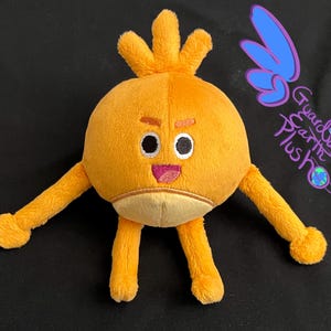 Puede incluir: Un juguete de peluche con forma de personaje redondo naranja con un peinado naranja puntiagudo, ojos negros, boca rosa y brazos y piernas largos de color naranja. El juguete est&aacute; sentado sobre una superficie negra.