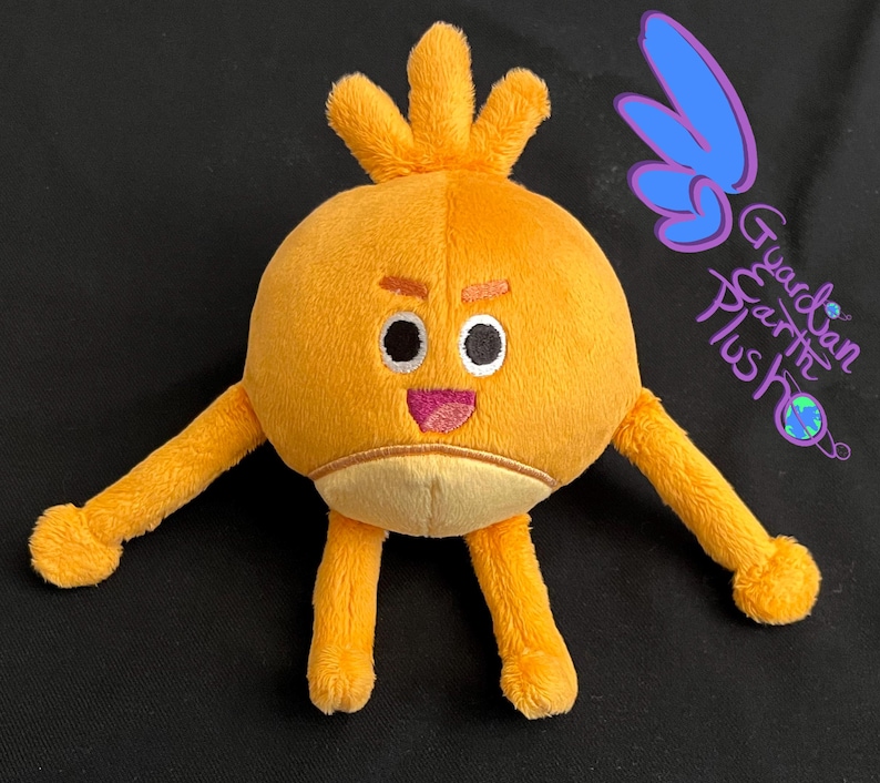 Puede incluir: Un juguete de peluche con forma de personaje redondo naranja con un peinado naranja puntiagudo, ojos negros, boca rosa y brazos y piernas largos de color naranja. El juguete est&aacute; sentado sobre una superficie negra.