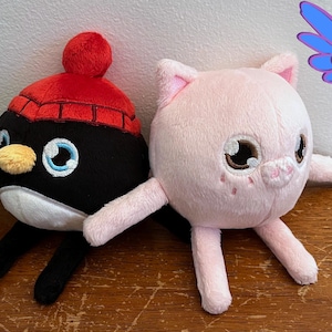 Puede incluir: Dos peluches, un ping&uuml;ino blanco y negro con un gorro rojo y un cerdo rosa con ojos marrones. Los juguetes est&aacute;n sentados sobre una superficie de madera. El texto "Guardian Earth Plush" es visible en el fondo.