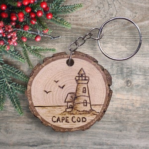 May include: A wooden keychain with a burnt image of a lighthouse and the words "Cape Cod" on a round piece of wood. The keychain is attached to a silver metal ring.