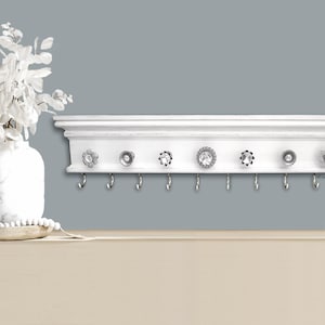 May include: White shelf with a decorative trim and eight hooks. The shelf has five crystal knobs with silver accents.