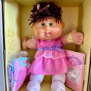 May include: A Cabbage Patch Kids doll in a box. The doll has brown hair, green eyes, and is wearing a pink dress with a purple bow. It is holding its thumbs up. Included are adoption papers and a set of clothes.