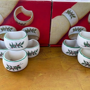 May include: A set of ceramic napkin rings and small bowls with a Christmas theme. The white rings and bowls have a green border and are decorated with green leaves and red berries. The set is displayed on a red and beige background.