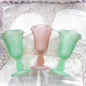 May include: Three frosted glass goblets in pastel colors: two green and one pink. The goblets have a tulip-shaped bowl and a slender stem, resting on a flat base. They are arranged on a white, lace-trimmed surface.