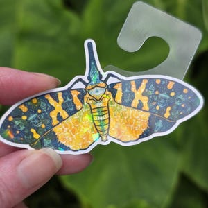 May include: A holographic sticker shaped like a lantern fly with a dark blue and yellow color palette. The wings feature intricate patterns of glowing lanterns and celestial motifs. The sticker includes a clear plastic tab at the top for hanging.