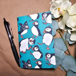 May include: A turquoise blue notebook with a repeating pattern of black and white puffins. The puffins are in various poses, some with their beaks open and some with their beaks closed. The notebook is a great way to add a touch of whimsy to your desk or bag.