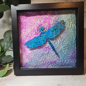 Puede incluir: Una pieza de arte mural enmarcada que presenta una libélula azul brillante con alas iridiscentes y patrones celestiales. La libélula está montada sobre un fondo texturizado con un degradado de tonos rosa, morado y azul en un marco cuadrado negro.