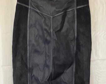 May include: A black pencil skirt with leather and suede panels. The skirt has a high waist and a knee-length hemline. Vertical seams and stitching details add structure. The skirt is hanging on a clear hanger.
