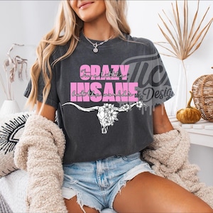 May include: Dark gray t-shirt with the text "Crazy, I'm a little insane but the world is ok" in pink. A white bull skull with floral accents is below the text. The model is wearing denim shorts and a cream-colored sweater.