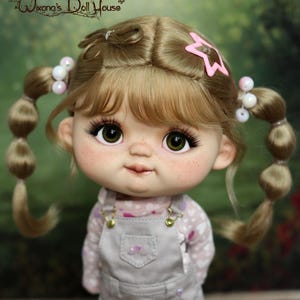 May include: A doll with blonde pigtails, decorated with pink star and pearl hair accessories. The doll is dressed in a light pink patterned shirt, grey dungarees, striped socks, and pink shoes. The doll has green eyes and rosy cheeks.