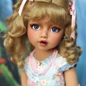 May include: A doll with blonde curly hair, blue eyes, and rosy cheeks. The doll wears a floral dress with pink accents and a pink beaded necklace. Pink ribbons adorn the doll's hair. The text "Wixana's Doll House" is visible.
