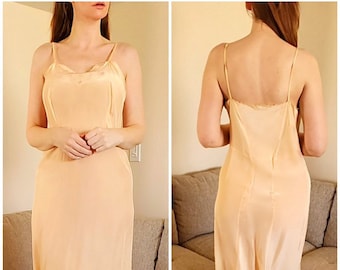 May include: A peach-colored slip dress with spaghetti straps. The dress has a subtle sheen and a delicate lace trim along the neckline. The dress falls to the mid-thigh and has a simple, elegant design.