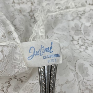 May include: Close-up of a white lace garment with a tag that reads "Intime CALIFORNIA SIZE S". The tag is held by a silver object. The lace has a delicate floral pattern.