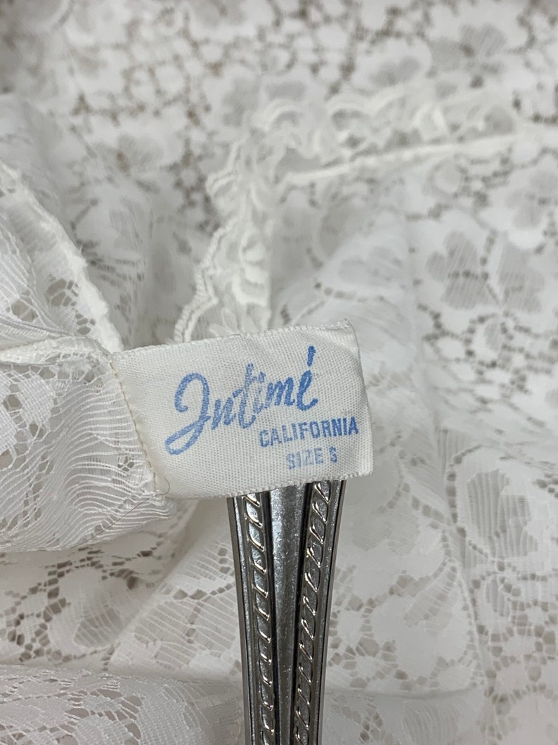 May include: Close-up of a white lace garment with a tag that reads "Intime CALIFORNIA SIZE S". The tag is held by a silver object. The lace has a delicate floral pattern.