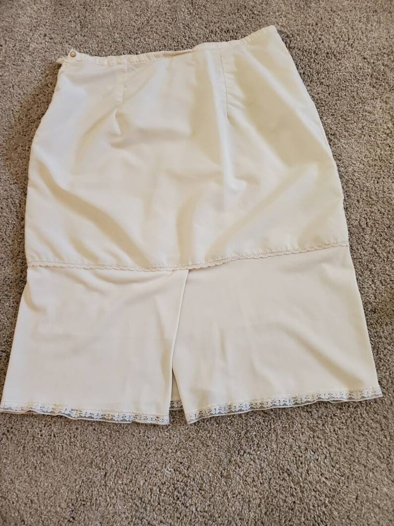 Pu&ograve; includere: Gonna vintage color crema con spacco sull'orlo. La gonna ha una vita alta e una chiusura con bottoni. L'orlo &egrave; rifinito con un delicato pizzo. Il tessuto sembra essere un materiale liscio e leggero.