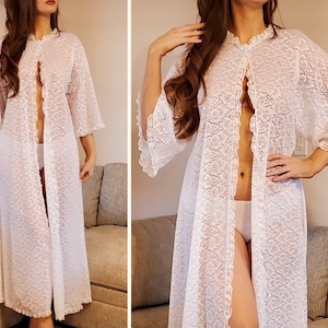 May include: A white lace robe with a floral pattern. The robe has a ruffled collar, wide sleeves, and a floor-length design. The robe is open in the front and has a delicate, feminine aesthetic.