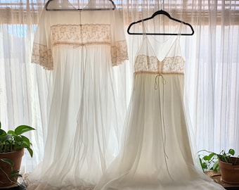 May include: Two vintage white lingerie pieces hang on black hangers. One is a long, sheer robe with lace trim and a tie closure. The other is a matching slip dress with spaghetti straps and lace detailing. Both are displayed in front of a window.