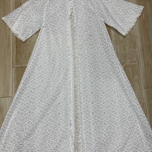 May include: A white, open-front lace robe with short sleeves and a delicate floral pattern. The robe features a scalloped trim along the edges and a small button closure at the neckline. The robe is made of a lightweight, semi-sheer fabric.