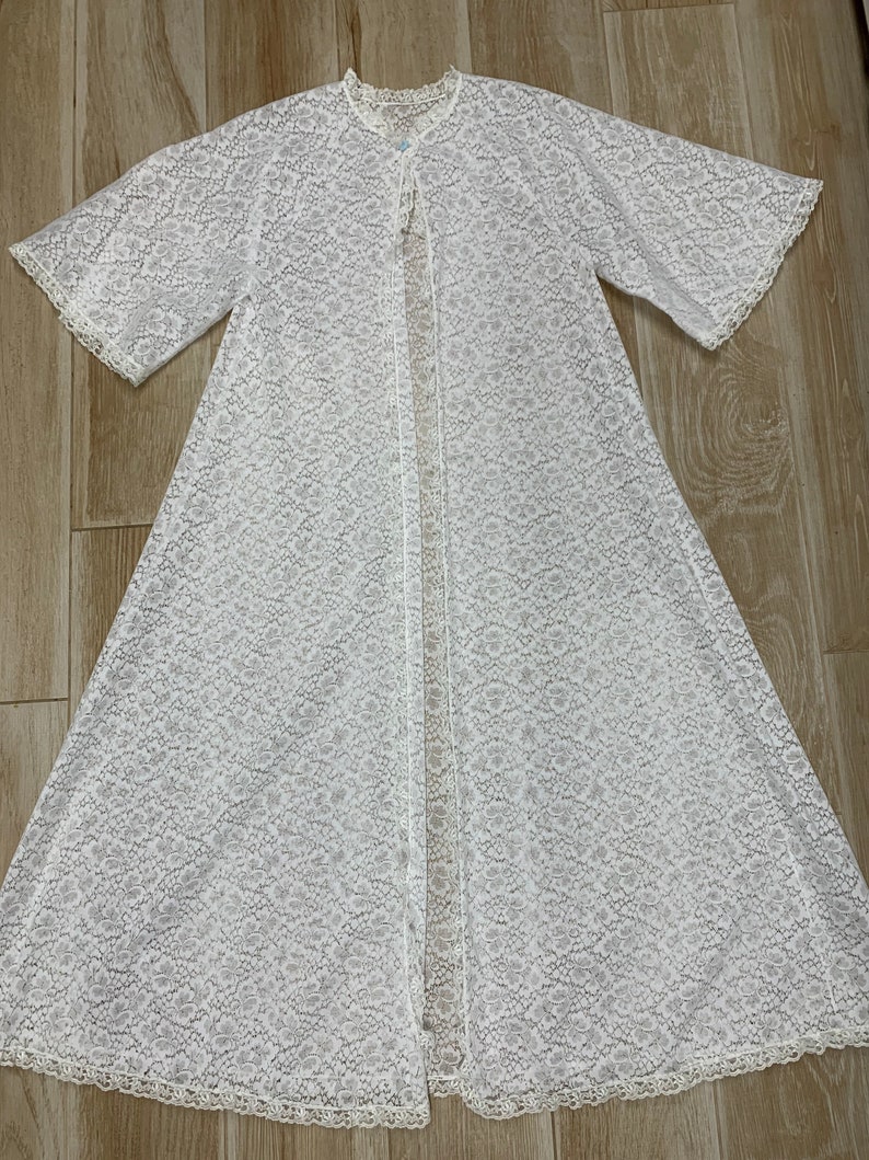 May include: A white, open-front lace robe with short sleeves and a delicate floral pattern. The robe features a scalloped trim along the edges and a small button closure at the neckline. The robe is made of a lightweight, semi-sheer fabric.