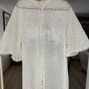 May include: A white lace robe with a floral pattern, long sleeves, and a button-up front. The robe has a ruffled collar and sleeve edges. The garment is hanging on a black hanger.