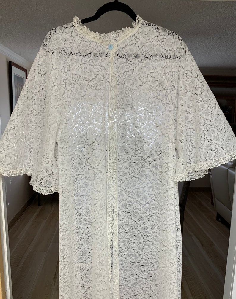 May include: A white lace robe with a floral pattern, long sleeves, and a button-up front. The robe has a ruffled collar and sleeve edges. The garment is hanging on a black hanger.