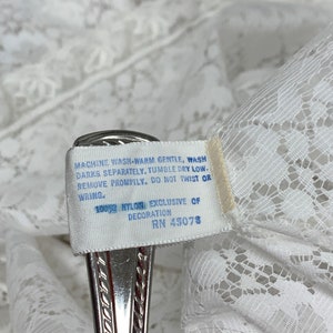 May include: Close-up of white lace fabric with a tag attached. The tag reads "MACHINE WASH WARM GENTLE, WASH DARKS SEPARATELY, TUMBLE DRY LOW-REMOVE PROMPTLY. DO NOT TWIST OR WRING. 100% NYLON EXCLUSIVE OF DECORATION RN 45078."