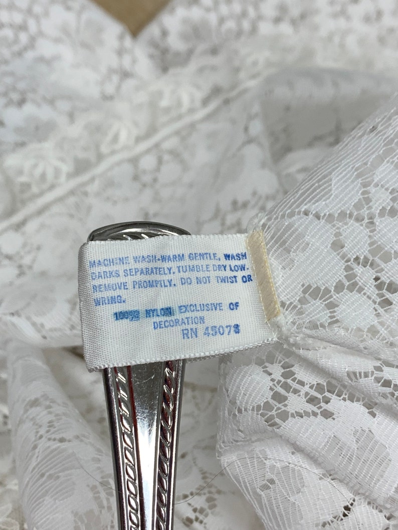 May include: Close-up of white lace fabric with a tag attached. The tag reads "MACHINE WASH WARM GENTLE, WASH DARKS SEPARATELY, TUMBLE DRY LOW-REMOVE PROMPTLY. DO NOT TWIST OR WRING. 100% NYLON EXCLUSIVE OF DECORATION RN 45078."