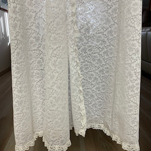 May include: A floor-length, off-white lace garment with a floral pattern. The garment has a button-up front and a scalloped lace trim along the hem and edges. The lace fabric is delicate and semi-sheer, creating a flowing, elegant look.