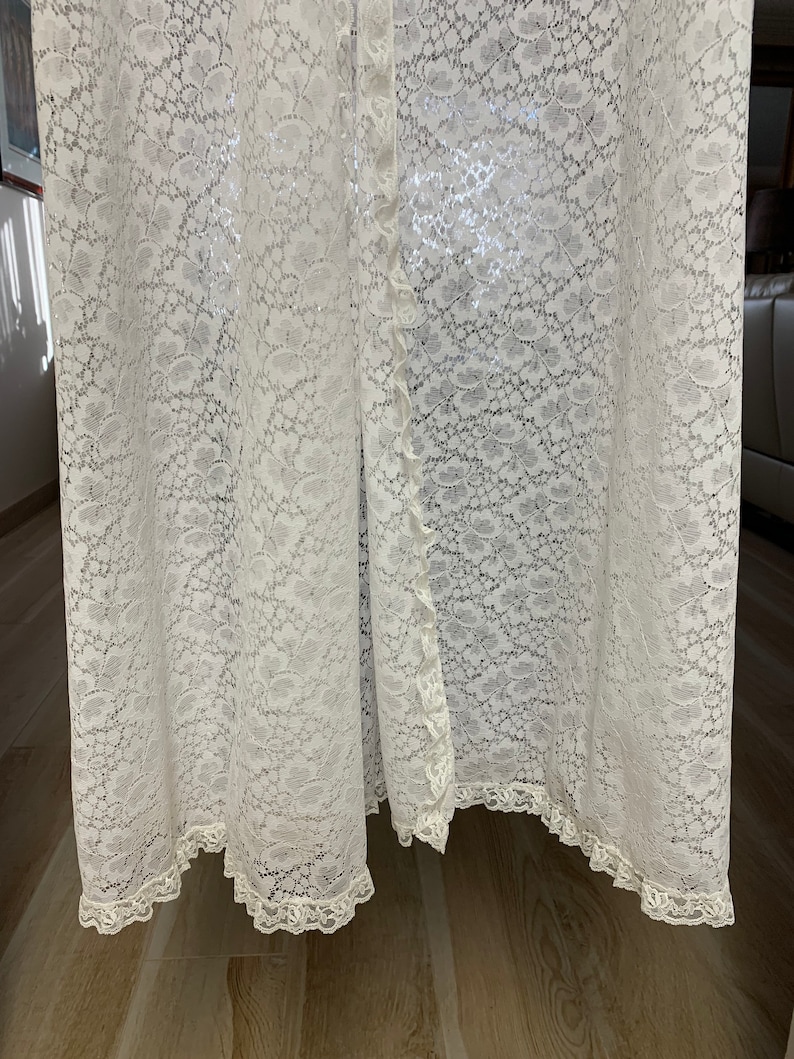 May include: A floor-length, off-white lace garment with a floral pattern. The garment has a button-up front and a scalloped lace trim along the hem and edges. The lace fabric is delicate and semi-sheer, creating a flowing, elegant look.