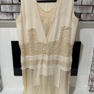 May include: Cream-colored vintage dress with a sleeveless design. The dress features intricate lace detailing on the bodice and skirt, with a layered skirt. The dress is hanging on a black hanger against a neutral background.