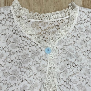 May include: A white lace garment with a delicate floral pattern. The garment features a ruffled collar and a row of small buttons down the front. A single light blue button is visible. The lace is intricately detailed.