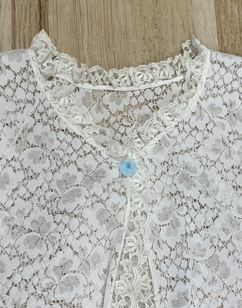 May include: A white lace garment with a delicate floral pattern. The garment features a ruffled collar and a row of small buttons down the front. A single light blue button is visible. The lace is intricately detailed.
