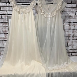 May include: Two vintage ivory-colored nightgowns hanging on black hangers. Both gowns feature lace trim around the neckline and sleeves, with delicate ribbon accents. The gowns are made of a flowing, lightweight fabric.