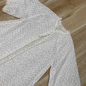May include: A white, floral lace robe with long, bell sleeves. The robe has a button closure at the neckline and delicate lace trim along the edges. The fabric is semi-sheer, revealing the texture of the floral pattern.