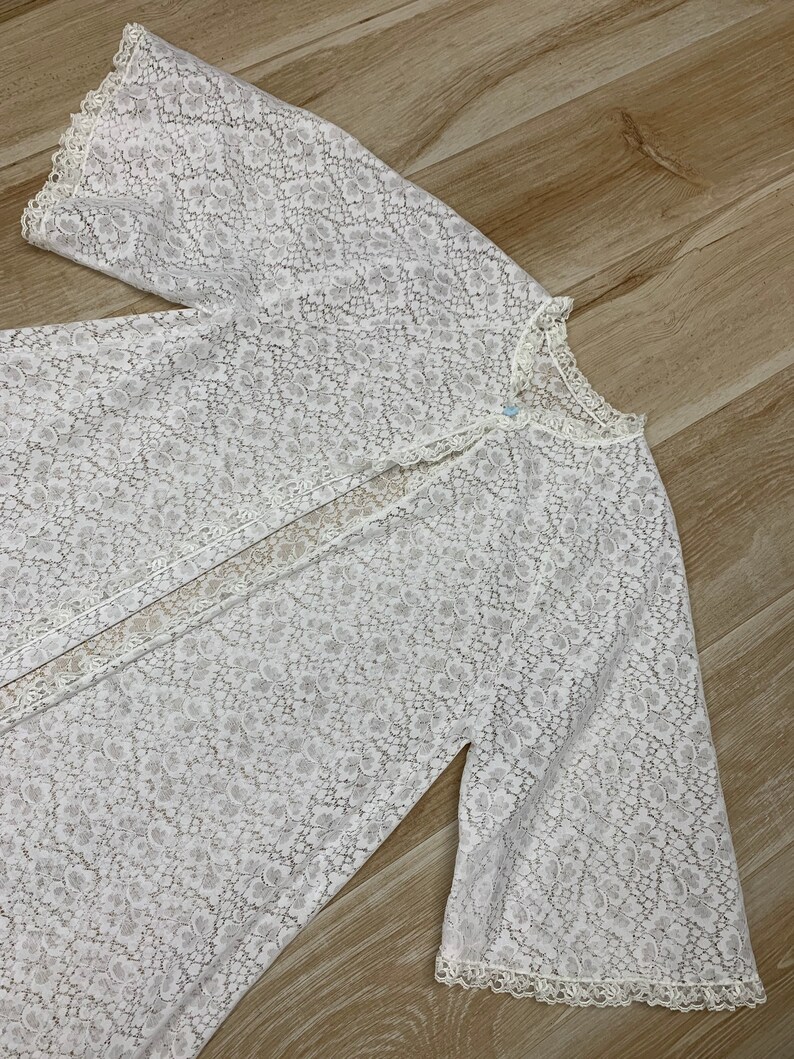May include: A white, floral lace robe with long, bell sleeves. The robe has a button closure at the neckline and delicate lace trim along the edges. The fabric is semi-sheer, revealing the texture of the floral pattern.