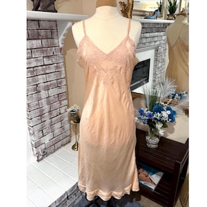 May include: A peach-colored slip dress with delicate spaghetti straps and a V-neckline. The bodice features lace detailing, and the dress has a flowing, knee-length design. The slip is displayed on a mannequin.