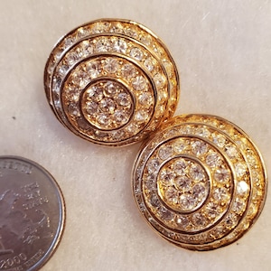 May include: Two gold-toned, round clip-on earrings with concentric circles of clear rhinestones. The earrings have a vintage look and are set against a light background. A quarter is next to the earrings.