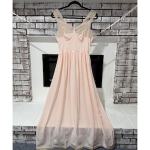 May include: A vintage blush-colored slip dress with a flowing silhouette. The dress features delicate lace detailing on the bodice and straps. It hangs on a black hanger against a brick wall background, showcasing its elegant design.