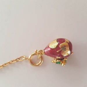 May include: A small, ornate pendant featuring a red, teardrop-shaped design with gold leaf accents. The pendant is attached to a gold chain and ring. A small green and gold accent is at the bottom of the pendant.