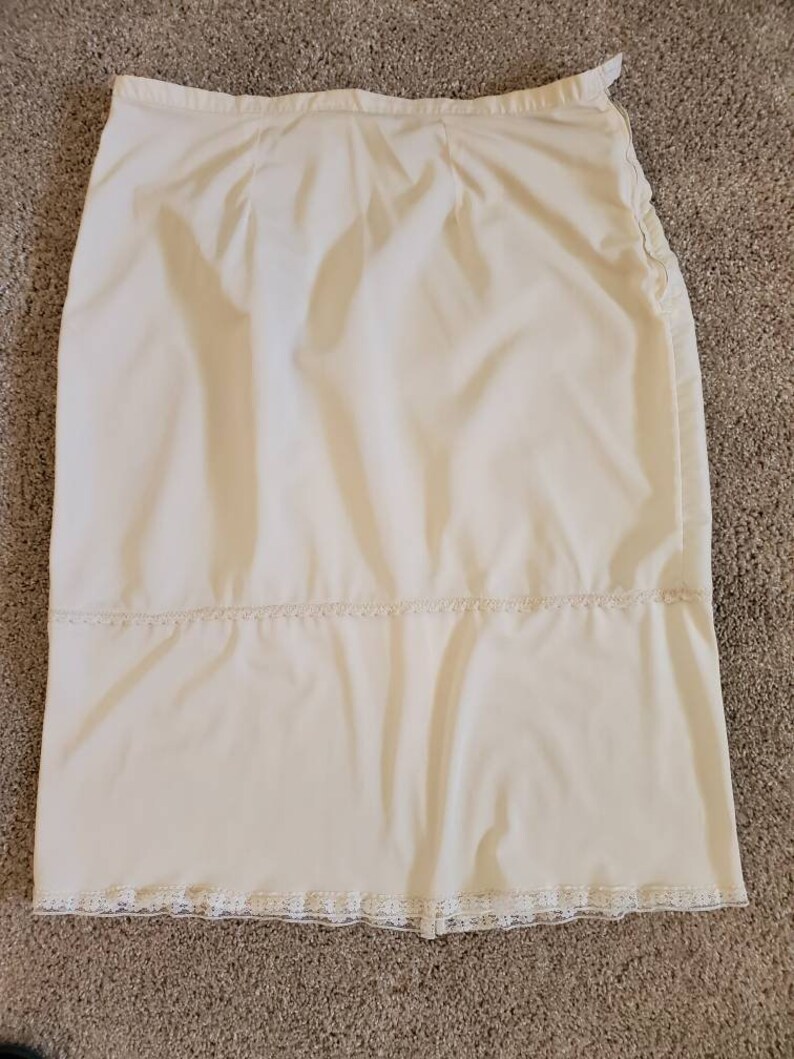 Pu&ograve; includere: Gonna color crema con taglio dritto, con delicati bordi in pizzo sull'orlo e una fascia orizzontale. La gonna ha una consistenza liscia, leggermente lucida e sembra essere realizzata in un tessuto leggero.