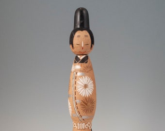 May include: A wooden doll with a black hat and a brown and white patterned kimono. The doll has a painted face with black hair and red lips.