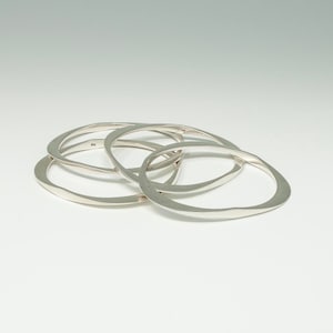 May include: Four silver bangles stacked on top of each other. The bangles are all smooth and have a slightly oval shape.